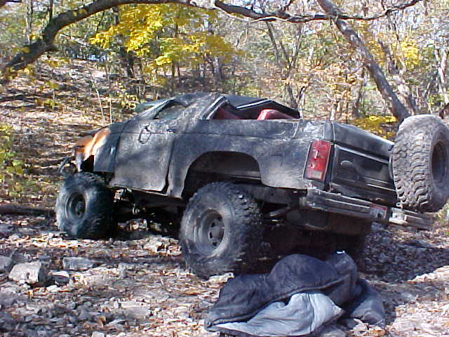 Stripped, mud-covered Ford Bronco trail rig parked on a rocky wooded trail