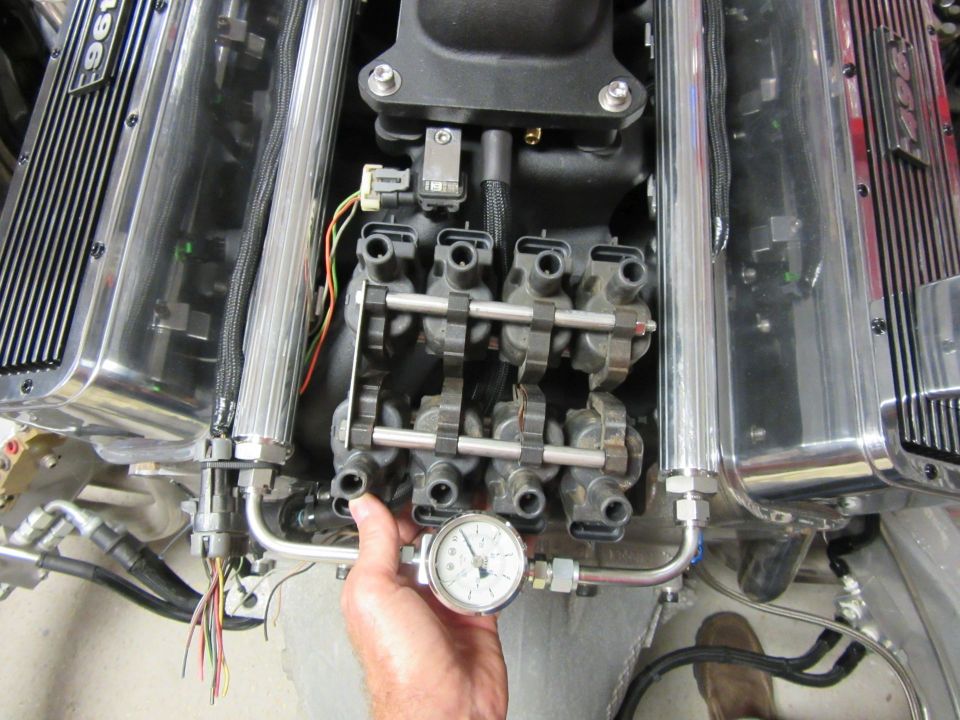 Close-up of a V8 engine fuel rail and pressure gauge. Polished valve covers have "196" visible.