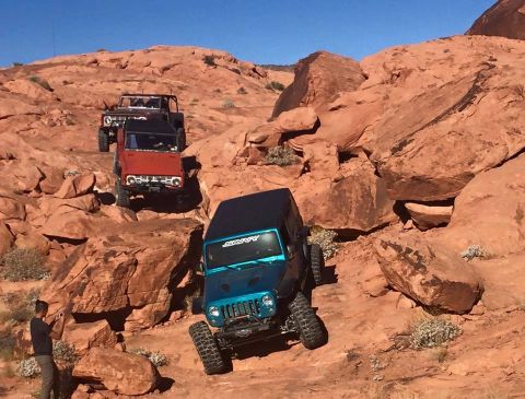 1979 Ford Bronco - A teal Jeep Wrangler is navigating a rocky, off-road trail, climbing over large red rocks. Two other off-road vehicles, including a red For…