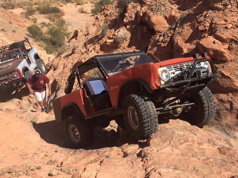 1979 Ford Bronco - A red, lifted 1979 Ford Bronco is navigating a rocky, off-road trail. Another white Bronco is visible further up the incline.
