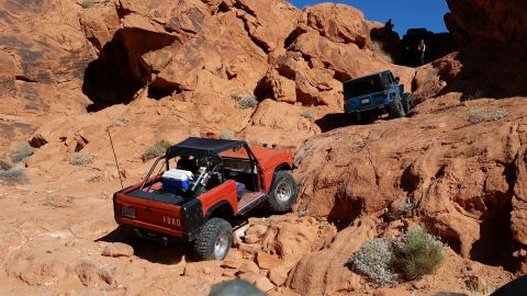 1979 Ford Bronco - A red 1979 Ford Bronco is navigating a rocky, off-road trail in a desert landscape with red rock formations. A blue Jeep Wrangler is also v…