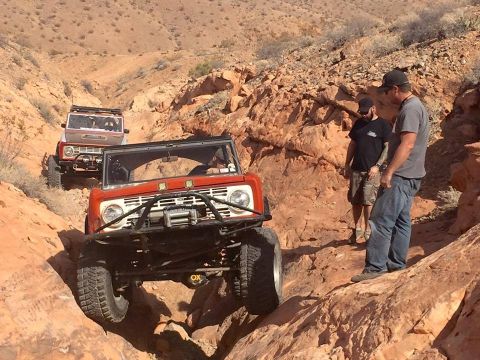 1979 Ford Bronco - Two orange and white Ford Broncos are navigating a rocky, off-road trail in a desert environment. Two men stand on the side of the trail, w…