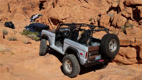 1979 Ford Bronco - A light gray Ford Bronco with a roll cage and off-road tires is parked on a rocky, desert terrain. Other off-road vehicles are visible in t…