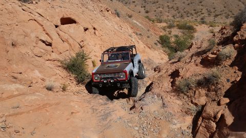1979 Ford Bronco - A modified light gray and brown 1979 Ford Bronco is navigating a rocky, off-road trail in a desert canyon. The vehicle features a roll cage…