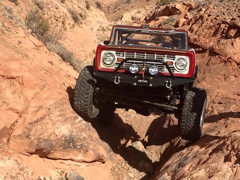 1979 Ford Bronco - A red 1970s Ford Bronco is navigating a rocky, off-road trail. The front of the vehicle is angled towards the camera, showcasing its large…