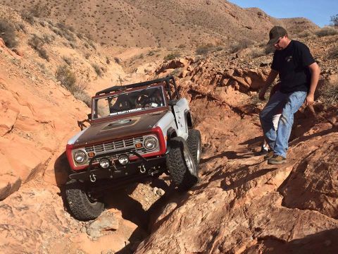 1979 Ford Bronco - A classic Ford Bronco is navigating a rocky, off-road trail with a person walking beside it. The Bronco is silver and brown with off-road t…