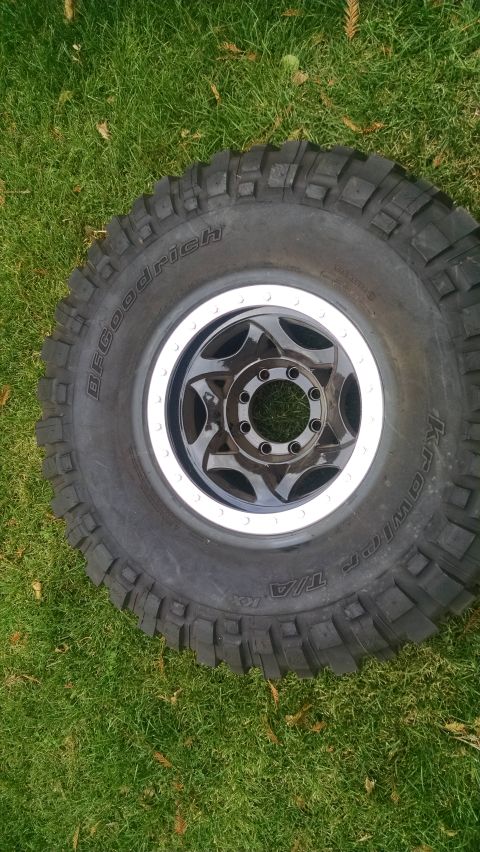 1985 Jeep CJ7 - A BFGoodrich All-Terrain T/A KO2 tire with a black multi-spoke rim is shown lying on a grassy lawn. The tire has aggressive tread blocks an…