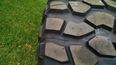 1985 Jeep CJ7 - A close-up, angled view of a large, aggressive off-road tire tread with visible metal studs embedded in the rubber blocks. The tire is rest…