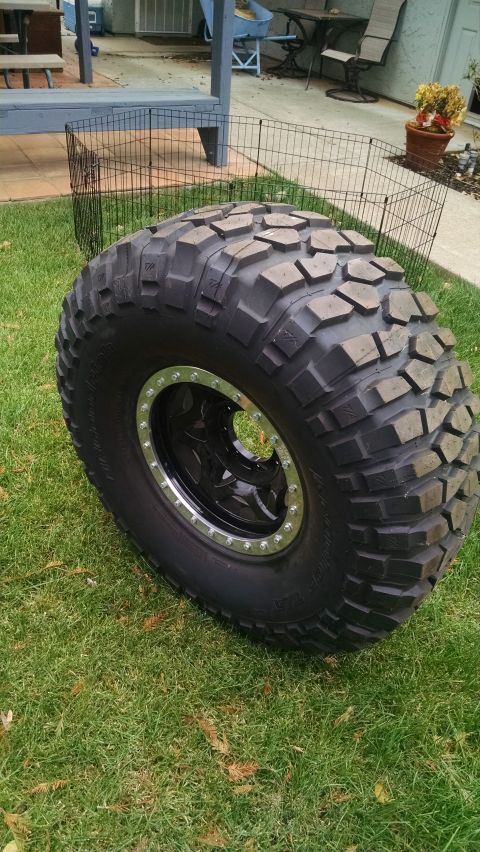 1985 Jeep CJ7 - A large, off-road tire with a black rim and silver beadlock is displayed on a grassy lawn. The tire has aggressive tread blocks and sidewal…