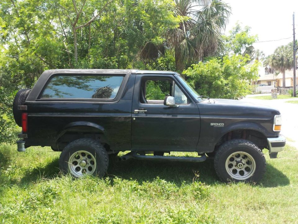 Black 1995 Ford Bronco parked in a grassy area with trees and houses.