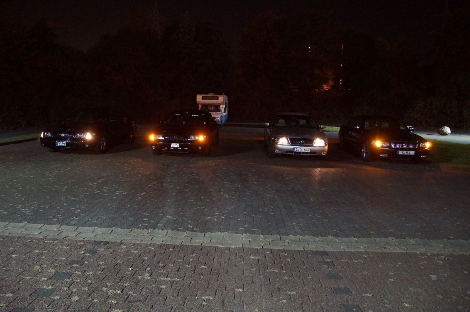 Four cars parked at night with headlights on, camper in background.