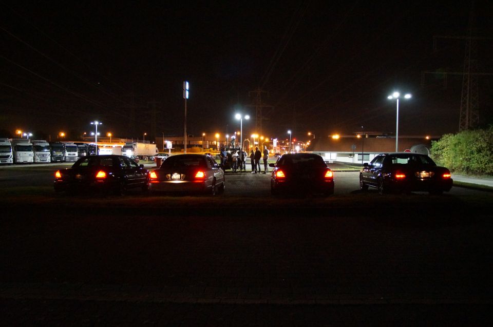 Four sedans parked at night with brake lights on, trucks in background.