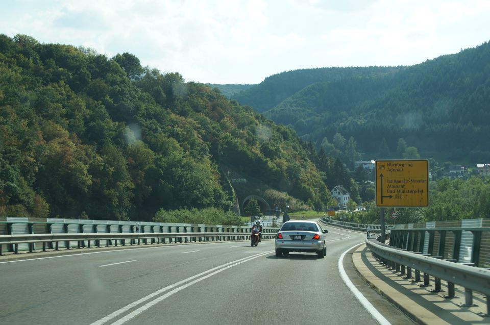 Silver sedan driving on a highway with a motorcycle ahead, surrounded by green hills and trees.