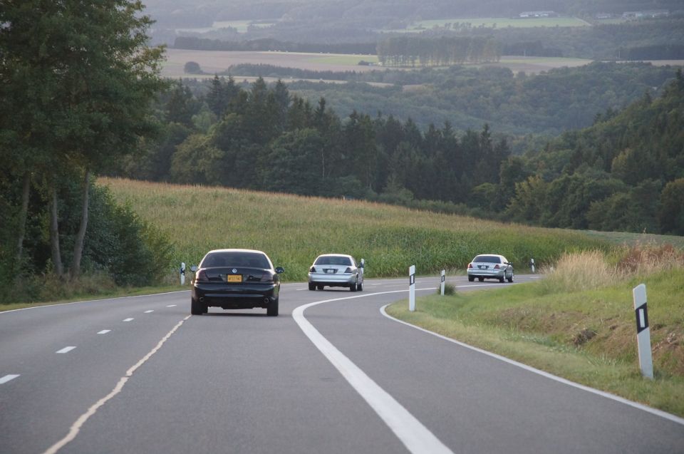 Two cars drive on a highway with trees and hills in the background