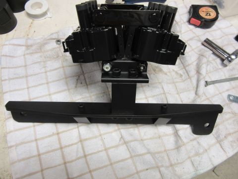 Black engine ignition coil pack assembly mounted on a bracket, with tools and tape measure nearby
