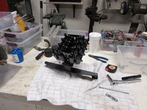 Black engine intake manifold with spark plug wires attached, resting on a checkered cloth on a workbench