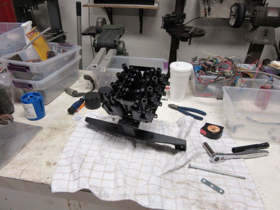 Black engine intake manifold with spark plug wires attached, resting on a checkered cloth on a workbench
