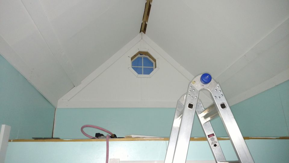 Interior of a room with light blue walls, white sloped ceiling, an octagonal window, and a silver extension ladder