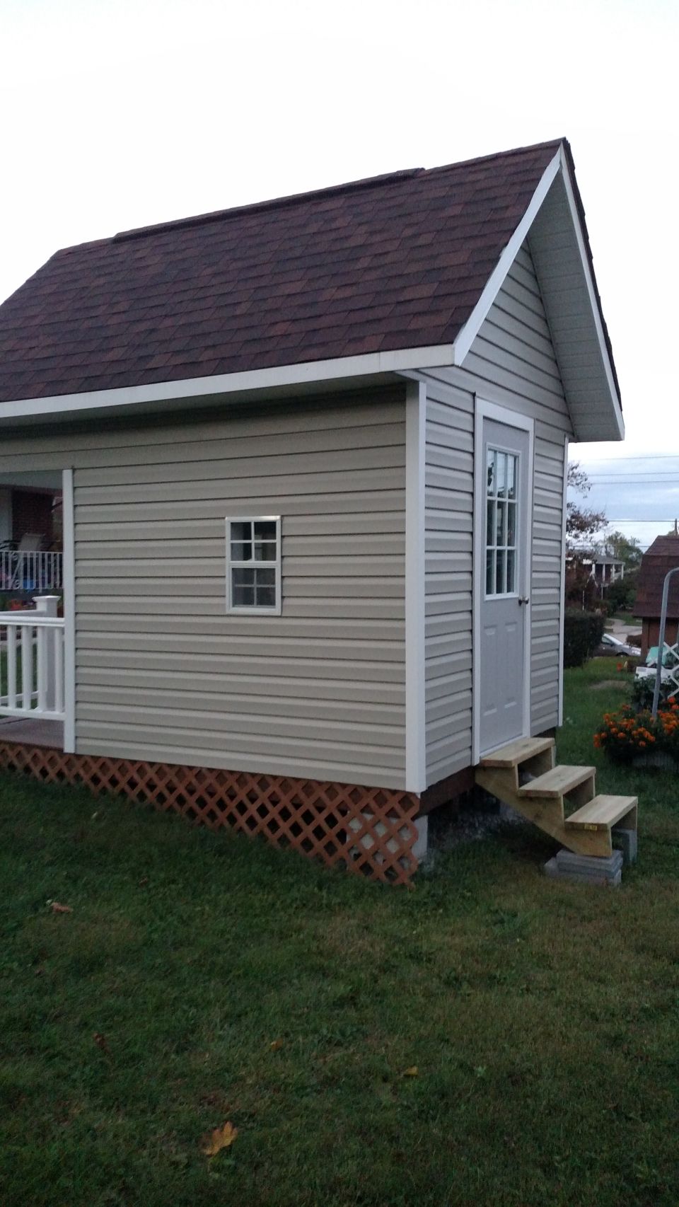 Tan vinyl-sided playhouse with a brown shingled roof and a white door with glass panes