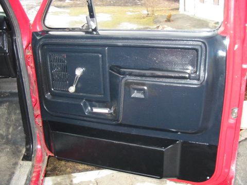 1981 Ford Bronco - The interior door panel of a red vehicle shows black trim and a manual window crank.
