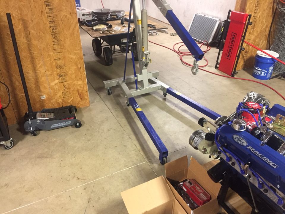 Blue Ford Racing engine with chrome accents, partially visible in a garage setting with an engine hoist and tools