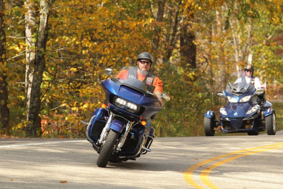 Blue motorcycle with a rider in an orange vest and helmet, leaning into a turn on a road with autumn trees. A blue three-wheeled vehicle follows behind