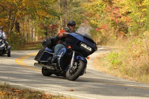 2014 Harley-Davidson FXDWGI Wide Glide - A blue motorcycle rides down a road lined with autumn foliage, with another vehicle visible in the background.