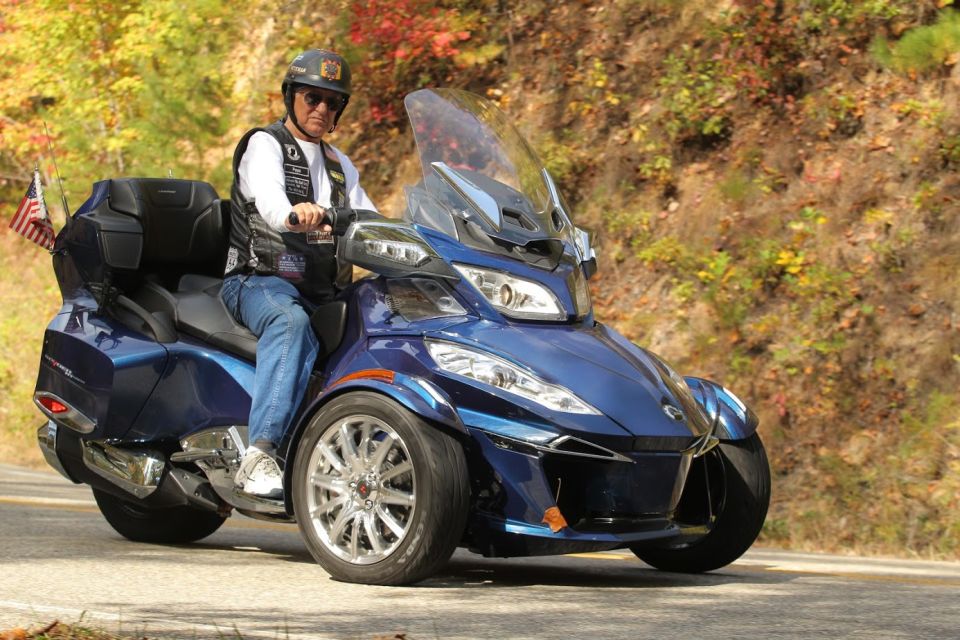 Blue three-wheeled Can-Am Spyder F3-S with a rider wearing a helmet and vest, driving on a road with trees in the background
