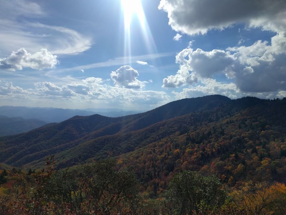 Mountain range under a bright, sunny sky with sunbeams breaking through clouds. Autumn foliage visible on the slopes
