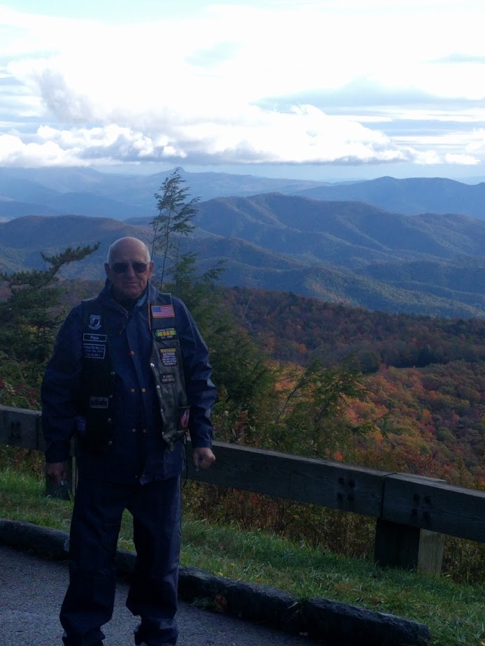 Man in a vest with patches stands in front of a mountain vista