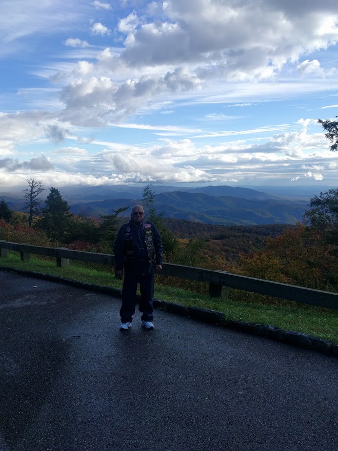 Man in a vest stands on a wet road overlooking a mountain vista under a cloudy sky