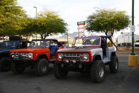 1979 Ford Bronco - Two modified Ford Broncos are parked side-by-side in a parking lot with trees and a highway in the background. The Bronco on the left is or…
