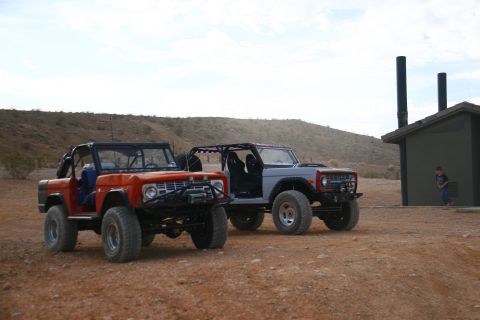 1979 Ford Bronco - Two modified Ford Broncos are parked on a dirt road in a desert landscape. The Bronco on the left is orange and the one on the right is gra…