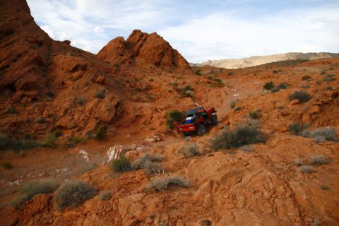 1979 Ford Bronco - A red off-road vehicle navigates a rocky, desert terrain with red rock formations and sparse vegetation under a cloudy sky.