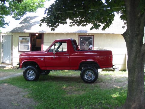 1981 Ford Bronco - A red Ford Bronco is parked on the grass in front of a small house, with a large tree to its right.