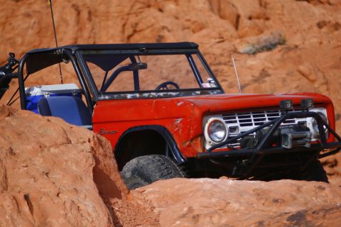 1979 Ford Bronco - A red 1979 Ford Bronco is shown off-road, navigating rocky terrain. The vehicle features a roll cage, a winch on the front bumper, and auxi…