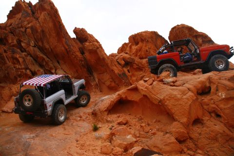 1979 Ford Bronco - Two classic Ford Broncos are shown off-roading on a rocky, red desert landscape. The Bronco on the left is silver with an American flag can…