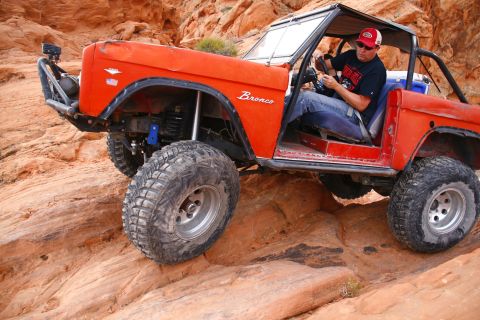 1979 Ford Bronco - An orange Ford Bronco is navigating a rocky, off-road trail with a driver at the wheel. The vehicle has large, knobby tires and a modified…
