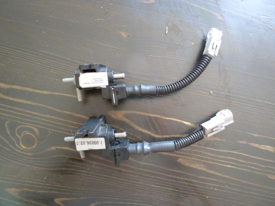 Two automotive solenoid valves with connectors and hoses on a wooden surface.