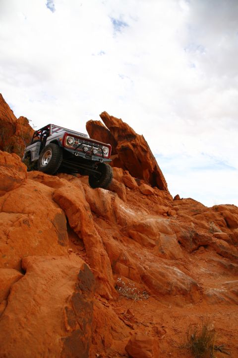 1979 Ford Bronco - A classic Ford Bronco is shown navigating a steep, rocky, orange-hued desert terrain under a cloudy sky. The off-road vehicle is angled upw…