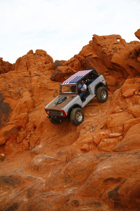 1979 Ford Bronco - A modified gray 1979 Ford Bronco is driving down a steep, rocky, orange desert trail. The vehicle has an American flag draped over its roll…