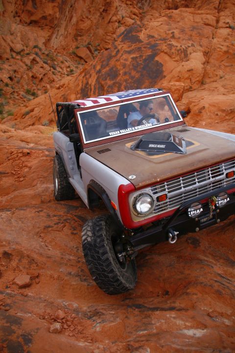 1979 Ford Bronco - A silver and brown 1979 Ford Bronco is driving off-road on a rocky, red terrain. The vehicle has an American flag decal on the roof and off…