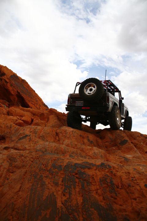 1979 Ford Bronco - A lifted 1979 Ford Bronco is shown from a low angle, climbing a steep, rocky, orange desert terrain under a cloudy sky. The rear of the SUV…