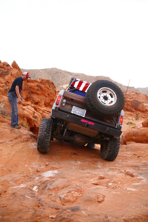 1979 Ford Bronco - A man in a red, white, and blue baseball cap and blue jeans stands on a rocky, desert terrain next to a brown Ford Bronco. The Bronco is an…