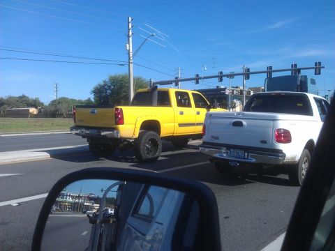 1994 Ford Bronco - A bright yellow pickup truck with large black rims is stopped at a traffic light behind a white pickup truck. The scene is viewed from the…