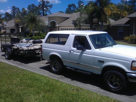 1994 Ford Bronco - A white 1990s Ford Bronco is parked on the side of a residential street, towing a utility trailer loaded with items. The vehicle is viewed…