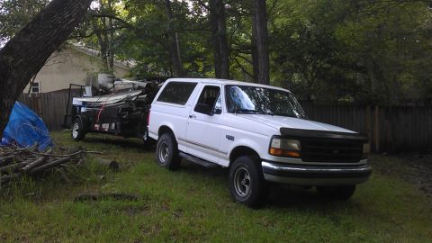 1994 Ford Bronco - A white 1990s Ford Bronco is parked in a grassy yard next to a trailer loaded with scrap metal. A large tree trunk is on the left side of t…