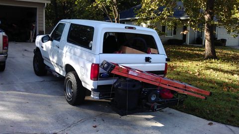 1994 Ford Bronco - A white Ford Bronco is parked in a driveway with a trailer attached to the rear. The trailer carries a large black trash can and an orange…