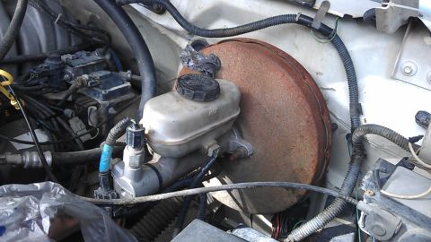1994 Ford Bronco - A close-up view of the brake master cylinder and brake booster assembly in a 1994 Ford Bronco engine bay. The brake booster is heavily rust…