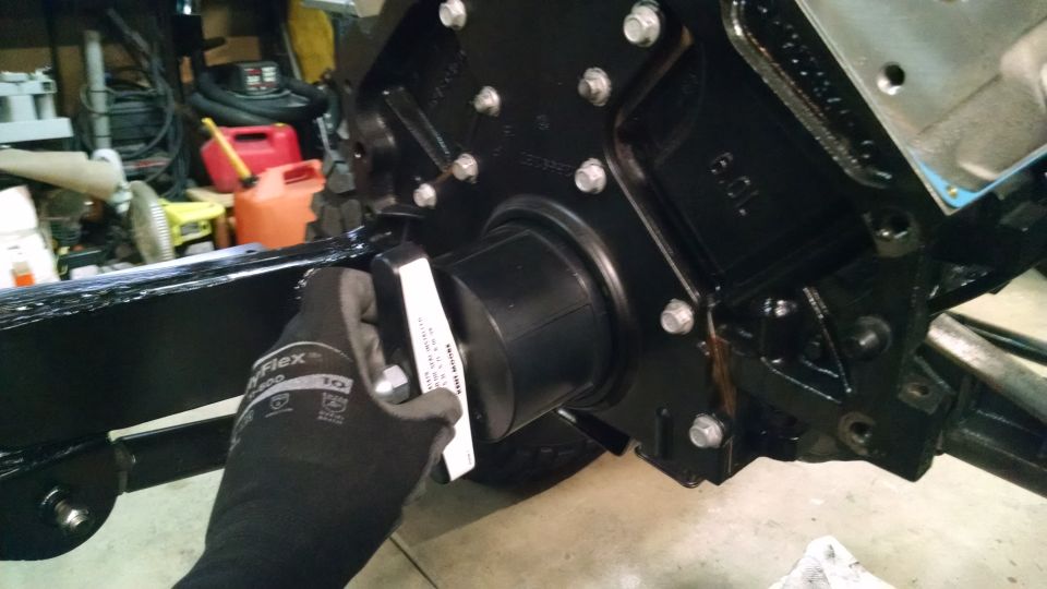 Gloved hand holding black part with white sticker against vehicle engine block and frame.